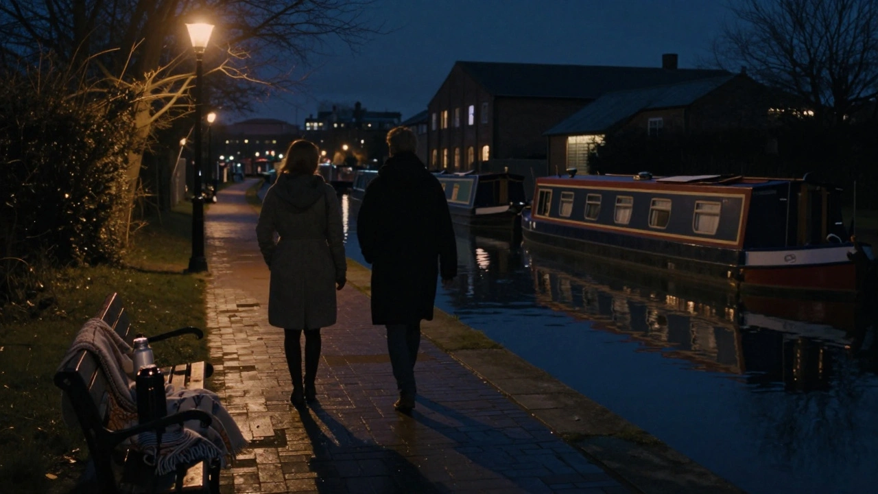 A couple strolls along Regent’s Canal at night, wrapped in coats, with a blanket and thermos, barges drifting in the dark water.